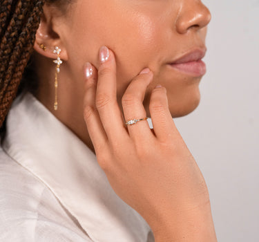 Woman wearing gold earrings and a ring, touching her face against a neutral background